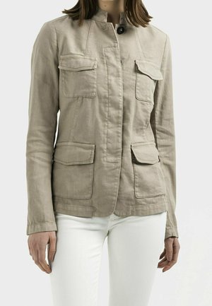 Mujer con chaqueta beige abotonada con cuatro bolsillos delanteros con solapa y pantalones blancos, vista desde los hombros hasta las caderas sobre un fondo liso.