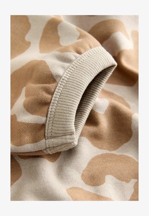 Tecido cu model de camuflaj prezintă pete de culoare bej și crem. Manseta are o textură ribbed, cu cusături clare pe margine.