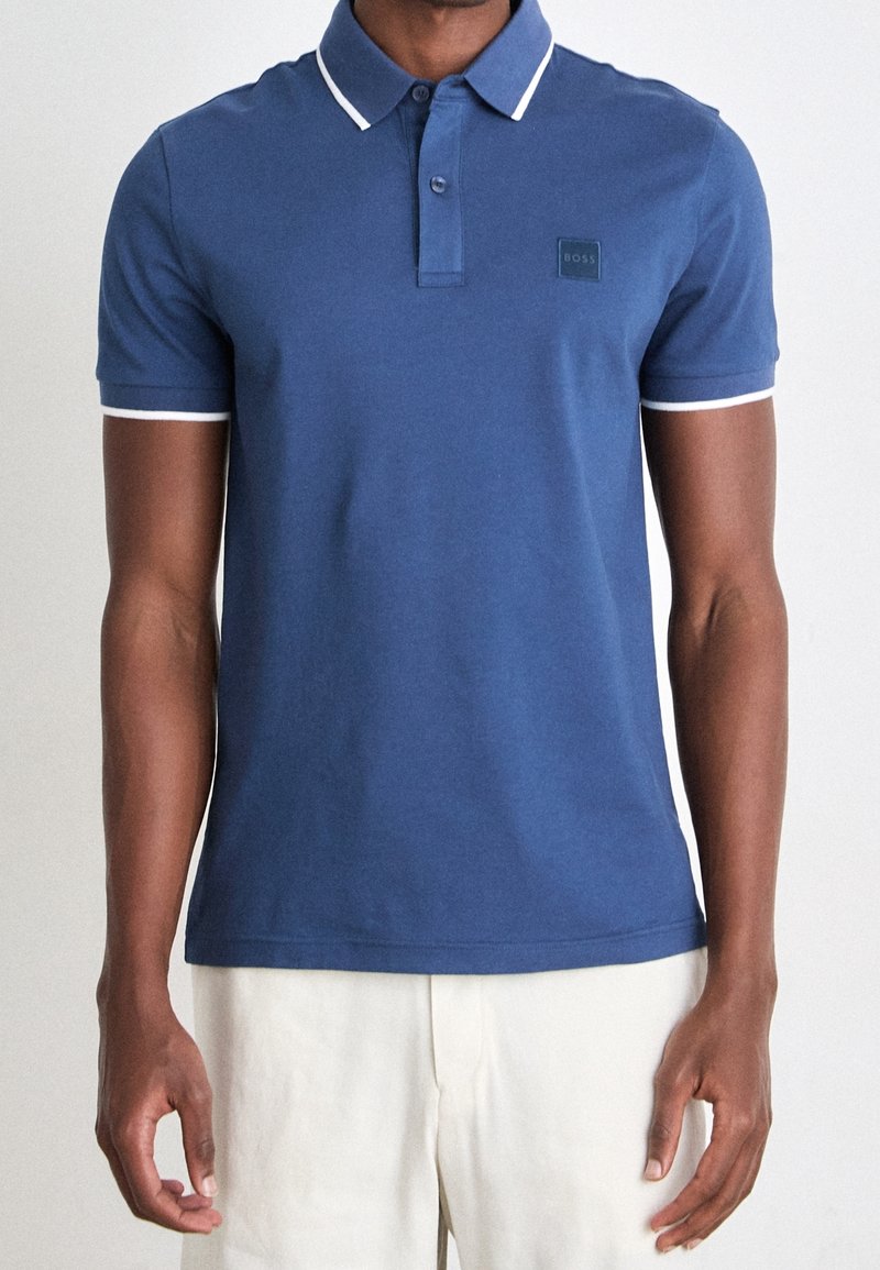 Homme portant un polo bleu à manches courtes avec une bordure blanche au col et aux manches, associé à un pantalon de couleur claire.