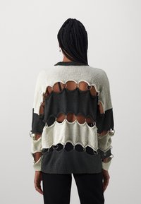Pull en tricot avec des sections ouvertes, comprenant des volumes noirs et blancs, des anneaux argentés reliant les couches. Tissu texturé avec une coupe ample.