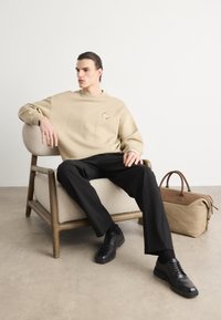 Beige kapucnis pulóver hímzett logóval, fekete nadrág és fekete cipő. Egy bézs táska fekszik a földön egy világos színű szék mellett.