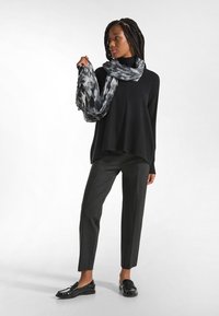 Maglione oversize nero con collo alto, abbinato a pantaloni grigi su misura e mocassini neri. Il modello indossa una sciarpa grigia con un motivo.