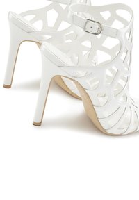 White high-heeled sandals with cut-out pattern and ankle strap. Glossy finish, pointed toe, and beige sole.