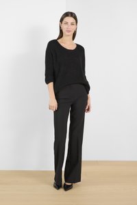 Sort strikket sweater med vid halsudskæring og trekvartærmer, parret med tætsiddende sorte bukser og spidse sorte hæle. Baggrund med trægulv.