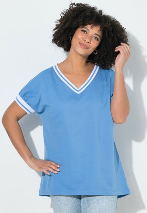 Smilende kvinde med krøllet hår iført en blå V-hals trøje med hvid stribet kant og lyseblå jeans, poserer med hånden på hoften.