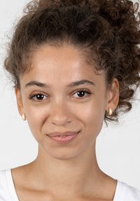 Guldtonade hoopörhängen med en texturerad yta, bars av en modell med lockigt hår, olivfärgad hudton och en enkel vit skjorta.