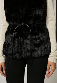 Musta tekoturkisliivi, jossa on teksturoitu pinta ja vyötäröllä musta nyöri. Yhdistetty kimaltavaan pitkähihnaiseen toppiin.