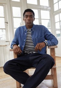 Camisa de ganga em azul claro com botões, sobreposta a uma t-shirt listrada em preto e branco. Calças azul-escuras e sapatilhas brancas. Sentado numa cadeira de madeira.
