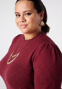 Dieprode gebreide trui met textuur in diamantenpatroon, ronde halslijn en pofmouwen. Gouden opvallende ketting gecombineerd met gouden oorringen.