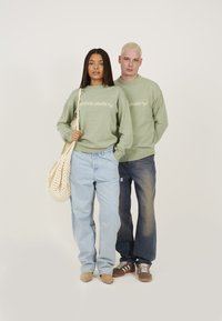 Grønne strikkede sweatshirts med "LEGACY" tekst, lys blå baggy jeans, beige sko og en beige hæklede taske. Enkel baggrund.