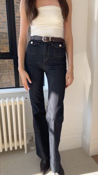 Jeans de ganga escuros de cintura alta com perna reta, apresentando bolsos frontais e um cinto texturizado castanho com uma fivela prateada.