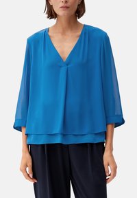 Femme portant un chemisier bleu ample à col en V superposé avec des manches 3/4 transparentes et un pantalon foncé sur un fond blanc uni.