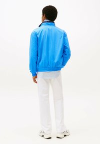 Blauwe lichtgewicht jas met elastische manchetten en zoom, gedragen over een lichtblauw gestreept shirt, gecombineerd met witte broek en sneakers.