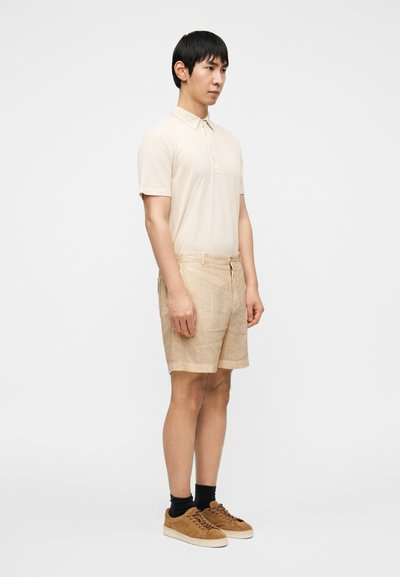 Μπεζ πόλο μπλούζα με γιακά, συνδυασμένη με ανοιχτό μπεζ λινά σορτς. Καφέ σουέτ αθλητικά παπούτσια με μαύρες κάλτσες. Απλή και καθημερινή εμφάνιση.