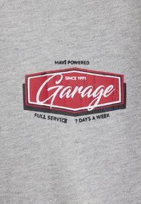Grauer Kapuzenpullover mit einem roten und schwarzen Logo, das den Text "Garage", gegründet 1991, und darunter "Vollservice 7 Tage die Woche" zeigt.