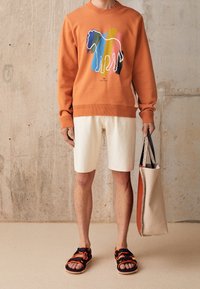 Sudadera naranja con un gráfico colorido de un caballo, combinada con pantalones cortos en un tono blanco roto y sandalias negras con acentos naranjas; sosteniendo un bolso de mano beige.