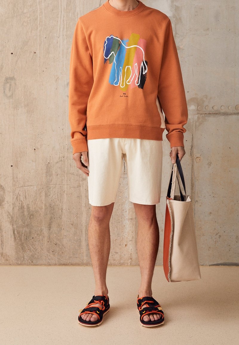 Sudadera naranja con un gráfico colorido de un caballo, combinada con pantalones cortos en un tono blanco roto y sandalias negras con acentos naranjas; sosteniendo un bolso de mano beige.