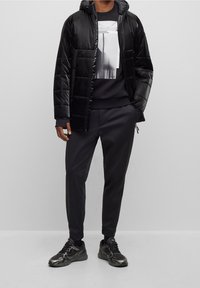 Černá zateplená bunda s kapucí, doplněná černou grafickou mikinou a odpovídajícími černými kalhotami. Vyznačuje se texturovanou tkaninou a elegantními sportovními botami.