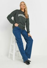 Felpa verde scuro con la scritta "MICHIGAN" stampata in grigio ad arco. Abbinata a jeans blu dritti e scarpe slip-on nere, il modello posa su uno sgabello.