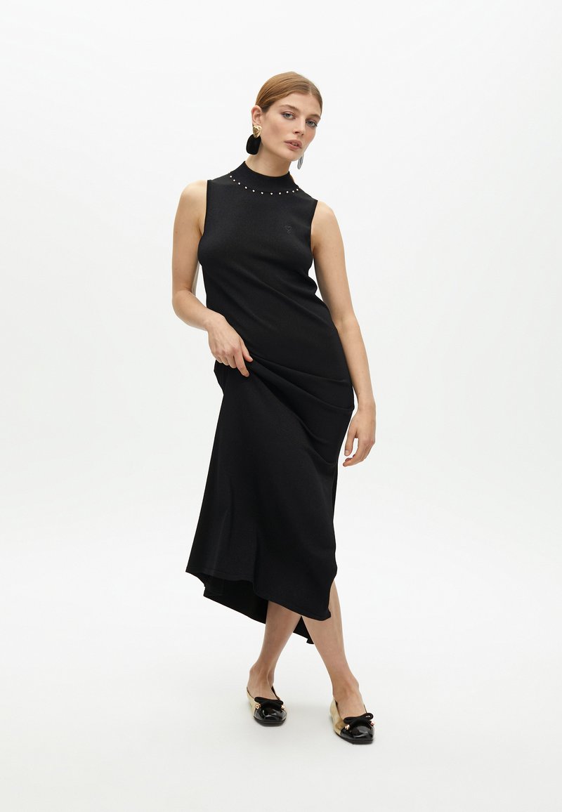 Mujer en un vestido midi negro sin mangas con escote tachonado y zapatos planos en negro y dorado posando frente a un fondo blanco.