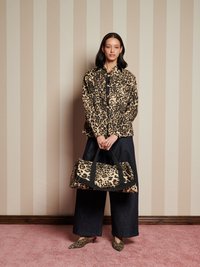 Mujer vestida con chaqueta de estampado de leopardo, zapatos a juego y sosteniendo un gran bolso de estampado de leopardo, de pie frente a una pared beige con rayas.