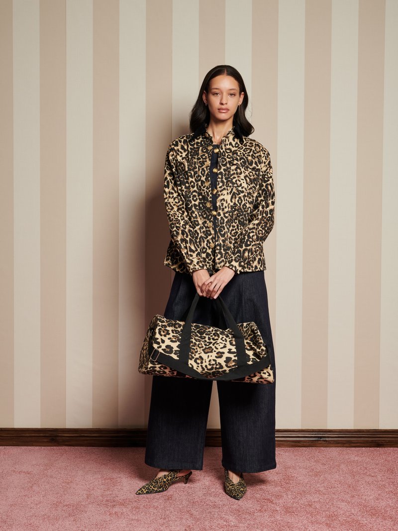 Mujer vestida con chaqueta de estampado de leopardo, zapatos a juego y sosteniendo un gran bolso de estampado de leopardo, de pie frente a una pared beige con rayas.
