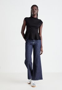 Woman standing against white background wearing black sleeveless peplum top, dark wide-leg jeans, and white sneakers.