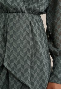 Zelena haljina s teksturiranim zig zag uzorkom, s naglašenim strukom i lepršavim materijalom. Dugi rukavi s prozirnim detaljima.