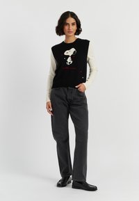 Gebreide gilet met een zwarte voorkant met Snoopy-print en rode accenten. De mouwen zijn crème. Gecombineerd met een grijze wijde broek en zwarte schoenen.