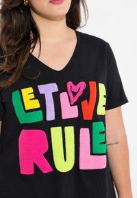 Sort V-hals T-shirt med flerfarvet, tekstureret skrift, der står "LAD KÆRLIGHEDEN REGERE" i fede, legende skrifttyper.