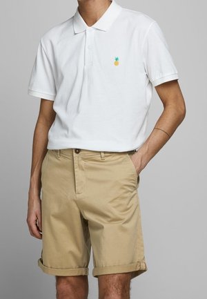 Witte poloshirt met een klein groen en oranje ananasembleem, gecombineerd met beige opgerolde shorts. Katoenen stof, casual pasvorm.