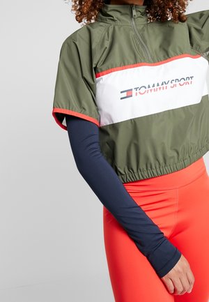 Kobieta w oliwkowo-zielonej krótkiej kurtce z logo "Tommy Sport", pod spodem granatowa bluzka z długim rękawem oraz jaskrawo pomarańczowe legginsy.