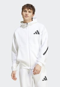 Veste à zip blanche avec logo Adidas noir, dotée d'une capuche avec cordon de serrage, d'une poche kangourou et de poignets côtelés. Texture lisse.
