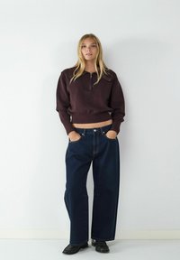 Brauner, mit Reißverschluss versehenen Pullover mit gerippten Bündchen; kombiniert mit lockeren, dunkelblauen Jeans mit vorderen Taschen und sichtbaren Nähten; schwarze Schuhe.