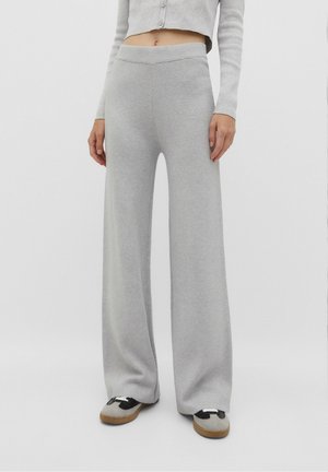 Frau trägt hellgraue, weit geschnittene Strickhosen, ein gekürztes, geknöpftes Strickjäckchen und zweifarbige Sneakers, steht vor einem einfarbigen Hintergrund.