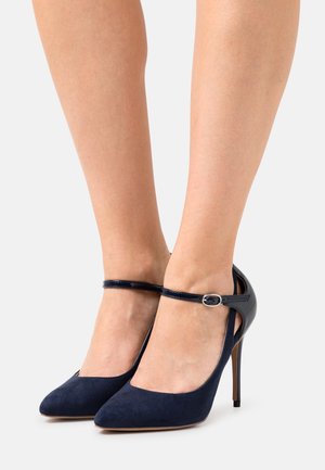 Talons hauts bleu marine avec une finition en suede et un bout pointu, dotés d'une bride de cheville en vernis brillant avec une boucle en argent.