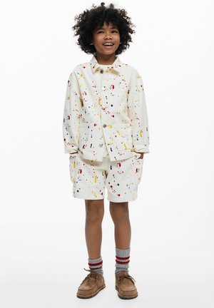 Fehér ruházat, rövid ujjú dzseki és rövidnadrág színes csepp mintával. Szürke zoknival és barna cipővel viselve.