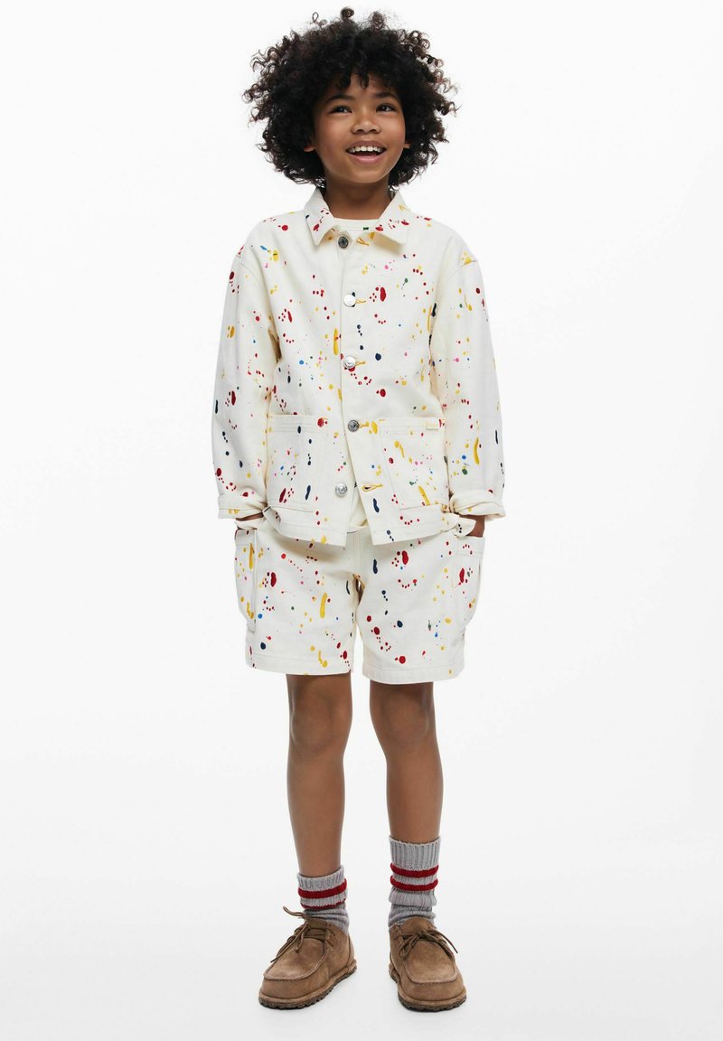 Ținută albă formată dintr-o jachetă cu mâneci scurte și pantaloni scurți cu un model stropit colorat. Poartă șosete gri și pantofi maro.