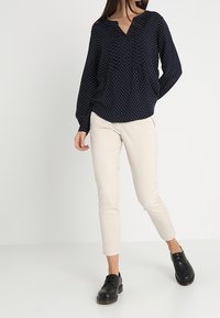 Námořnická halenka s bílými puntíky, výstřih do V, dlouhé rukávy, v kombinaci s béžovými slim-fit kalhotami a černými šněrovacími botami.