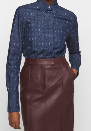 Donna che indossa una camicia di jeans blu scuro con fantasia e bottoni e una gonna di pelle marrone scuro a vita alta, con una mano in tasca.