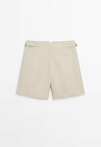 Shorts beige taille haute avec bretelles à boucle sur les côtés et deux poches passepoilées à l'arrière, présentés à plat sur un fond blanc.