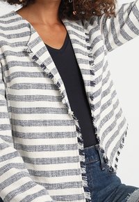 Giacca a righe bianca e blu navy, realizzata in tessuto testurizzato, presenta bordi sfrangiati e un design aperto nella parte frontale, abbinata a un top nero.