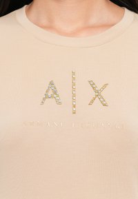Beige színű póló, melyen arany és ezüst strasszokkal díszített "A|X" logó található, alatt pedig a "ARMANI EXCHANGE" felirat van megjelenítve, azonos színben.