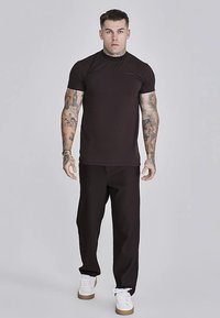 Temno rjava oprijeta majica s kratkimi rokavi, kombinirana s črnimi hlačami z rebrasto strukturo in belimi supergami s rjavimi podplati. Enostavna zasnova in gladka tekstura.