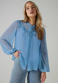 Blusa azul clara con tejido transparente y texturizado, manga larga, escote con volantes y cierre de botones en la parte delantera. Presenta un patrón de lunares.