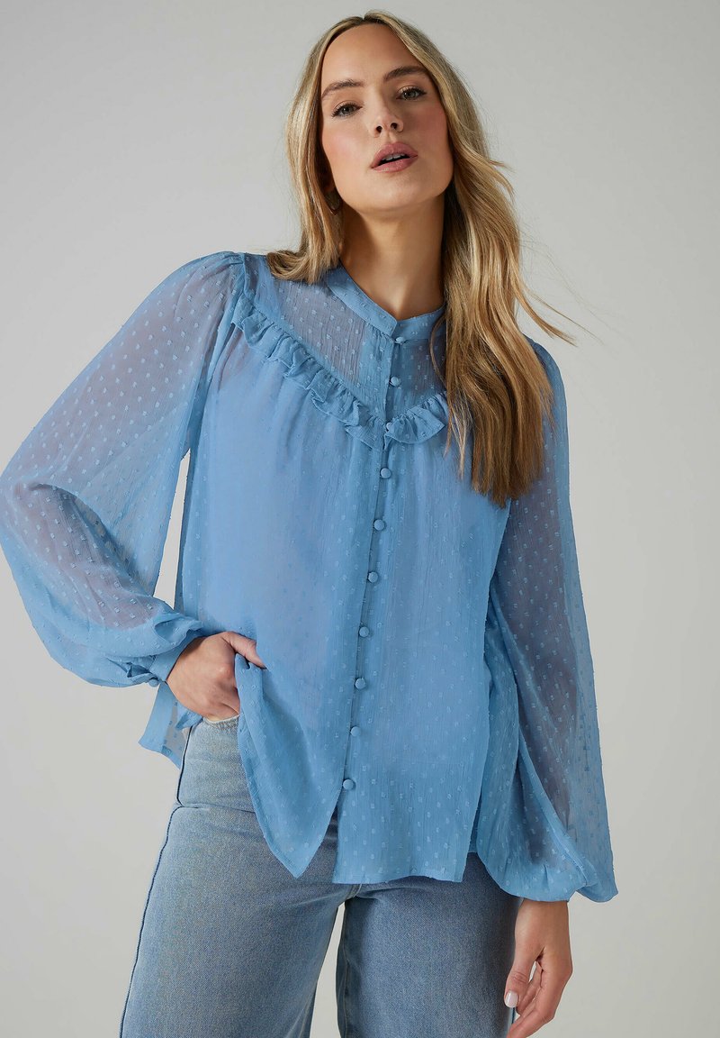 Blusa azul clara con tejido transparente y texturizado, manga larga, escote con volantes y cierre de botones en la parte delantera. Presenta un patrón de lunares.