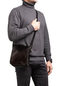 Borsa a spalla in pelle marrone scuro con un design piatto e rettangolare e una tracolla regolabile, tenuta da una persona che indossa un maglione a collo alto grigio.