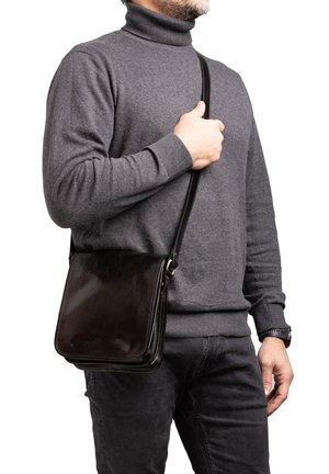 Borsa a spalla in pelle marrone scuro con un design piatto e rettangolare e una tracolla regolabile, tenuta da una persona che indossa un maglione a collo alto grigio.