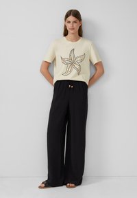 Beige T-Shirt mit einem bunten Seestern-Design, kombiniert mit lockeren schwarzen Hosen mit Kordelzug und Holzperlenakzenten.