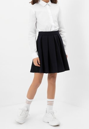Jeune personne portant une chemise blanche à col, une jupe plissée noire, des chaussettes blanches à trois rayures et des baskets blanches épaisses, debout devant un fond uni.